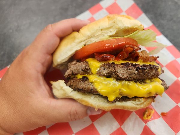 Double cheeseburger with bacon, lettuce and tomato on red-checkered paper, held in one hand