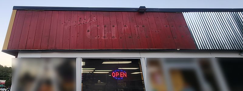 The building’s red vertical siding above the front windows, with the faded outline of the old Heidi’s sign letters and bolt holes still visible