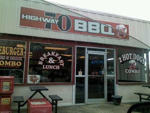 The Highway 70 BBQ storefront: a red sign band with a cartoon pig above a window wall lettered Breakfast and Lunch and 2 Hot Dogs Combo, a glass front door, and picnic tables out front