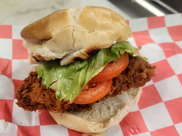 Chicken Cutlet Sandwich: crispy fried chicken cutlet with lettuce and tomato on a bun, on red-checkered paper
